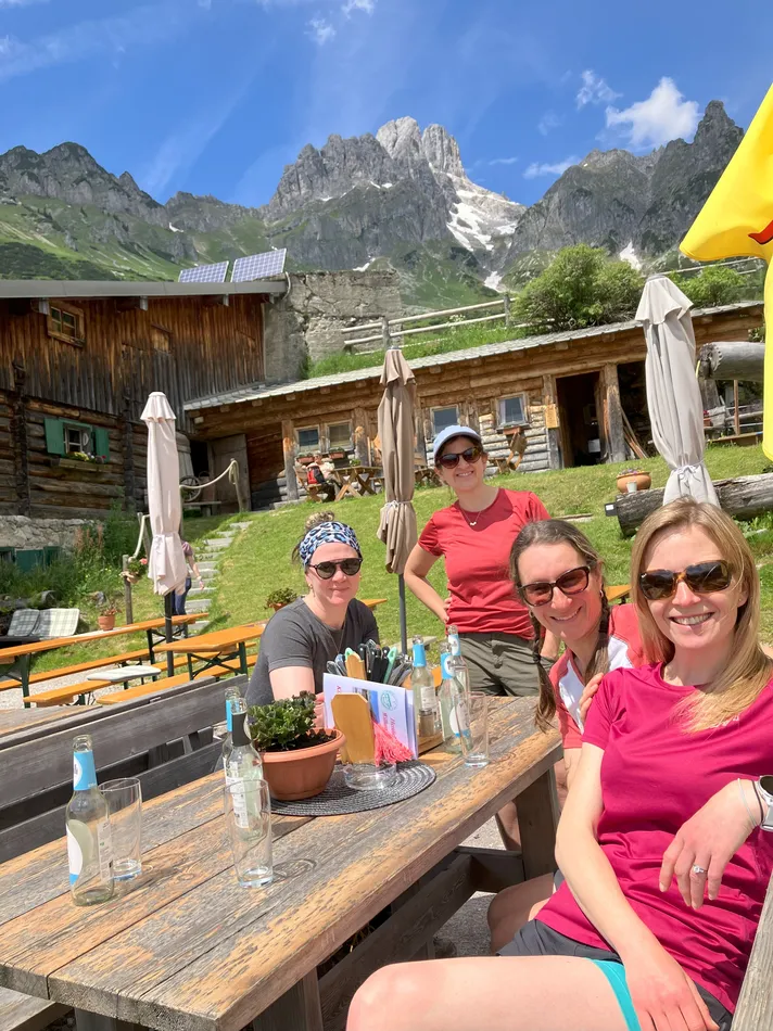 Four women relax at a rustic mountain hut table beneath dramatic jagged peaks and patches of lingering snow. Stopping for food and drinks between hikes makes hut trips one of the most social active girls trip ideas.