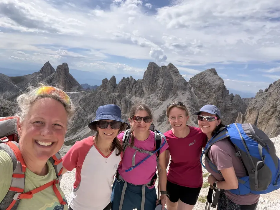 Five women wearing hiking packs smile together in front of dramatic jagged mountain peaks. Their group hike combines challenging alpine scenery with time spent traveling together.