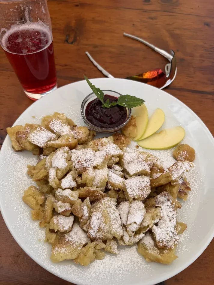 A plate of shredded pancake pieces is heavily dusted with powdered sugar and served with apple slices and a small bowl of dark berry sauce. A glass of red drink and a pair of sunglasses sit on the wooden table beside it.