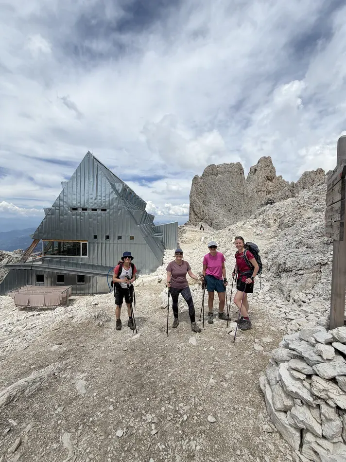 Four women with backpacks and trekking poles stand beside a modern metal mountain hut surrounded by pale rocky peaks. The remote hut provides a stopping point high in the mountains.