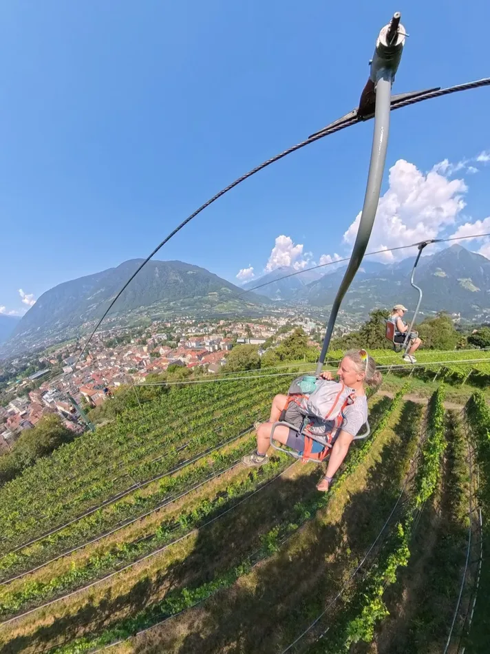 A woman wearing a hiking backpack rides an open chairlift above neat rows of hillside vineyards with the town and mountains below. The lift provides a scenic connection before or after sections of the 3 day Meraner Höhenweg hike.