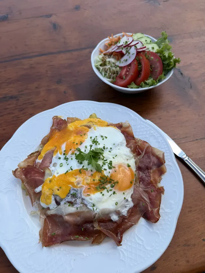 Two fried eggs topped with chopped herbs are served over thin slices of cured ham on a white plate. A small side salad contains lettuce tomatoes cucumber radish and shredded vegetables.