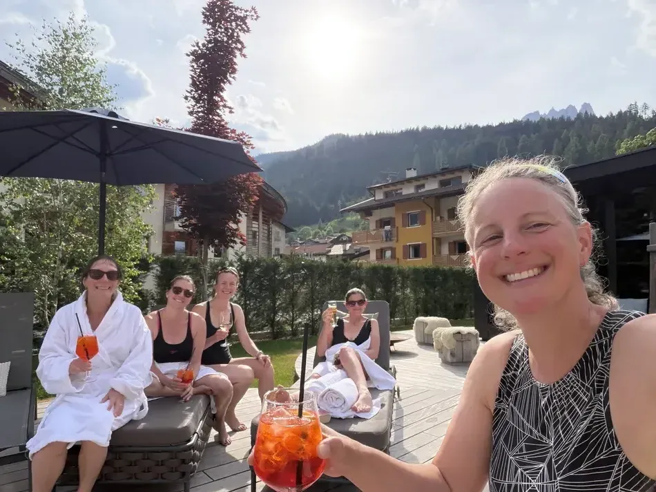 Five women relax on outdoor loungers in robes and swimsuits while holding orange drinks in the sunshine. A spa afternoon offers a relaxing contrast to hiking and other active girls trip ideas.